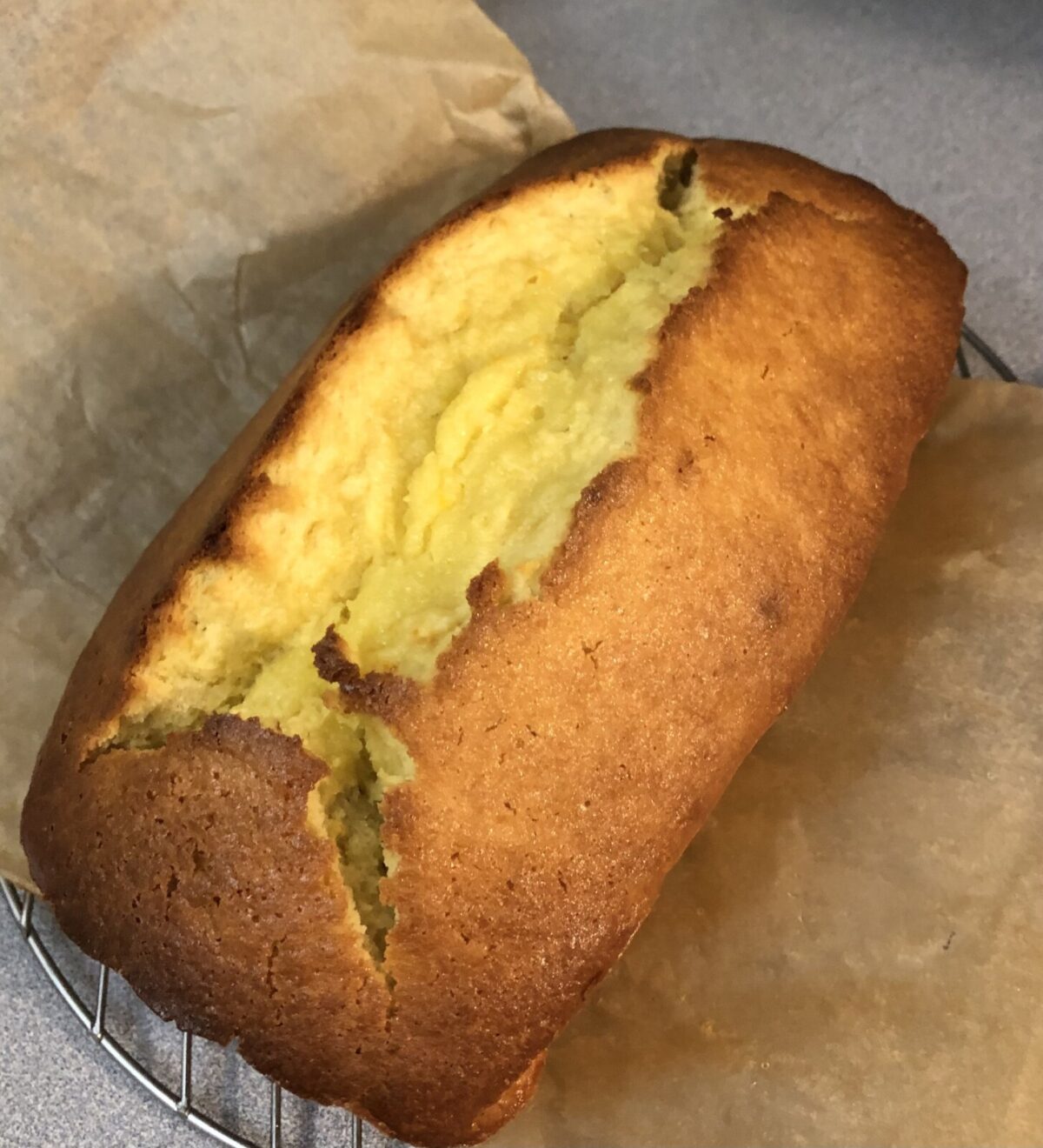 A golden-brown loaf of Whole Orange Blender Cake rests on parchment paper atop a cooling rack, its cracked top revealing a moist, citrusy yellow interior.