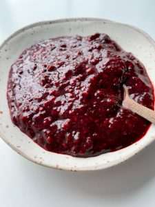 A close-up of a white, speckled ceramic bowl filled with thick, dark red raspberry chia jam. A small wooden spoon rests in the jam, and the bowl is set on a plain white surface.
