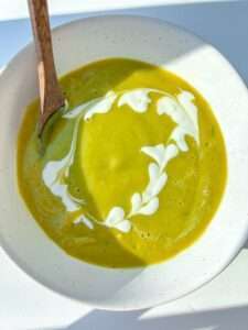 A bowl of Healthy Cream of Broccoli Soup with a swirl of white cream forming a heart-shaped pattern, served with a wooden spoon on the side. Bright natural light casts soft shadows across the bowl.