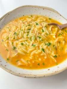 A bowl of Hidden Veggie Orzo Soup with a yellow broth, garnished with chopped herbs, and a wooden spoon resting inside the bowl.