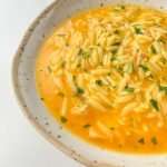 A close-up of Hidden Veggie Orzo Soup with a creamy orange broth, garnished with chopped herbs, served in a rustic, speckled ceramic bowl on a white surface.