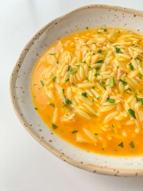 A close-up of Hidden Veggie Orzo Soup with a creamy orange broth, garnished with chopped herbs, served in a rustic, speckled ceramic bowl on a white surface.