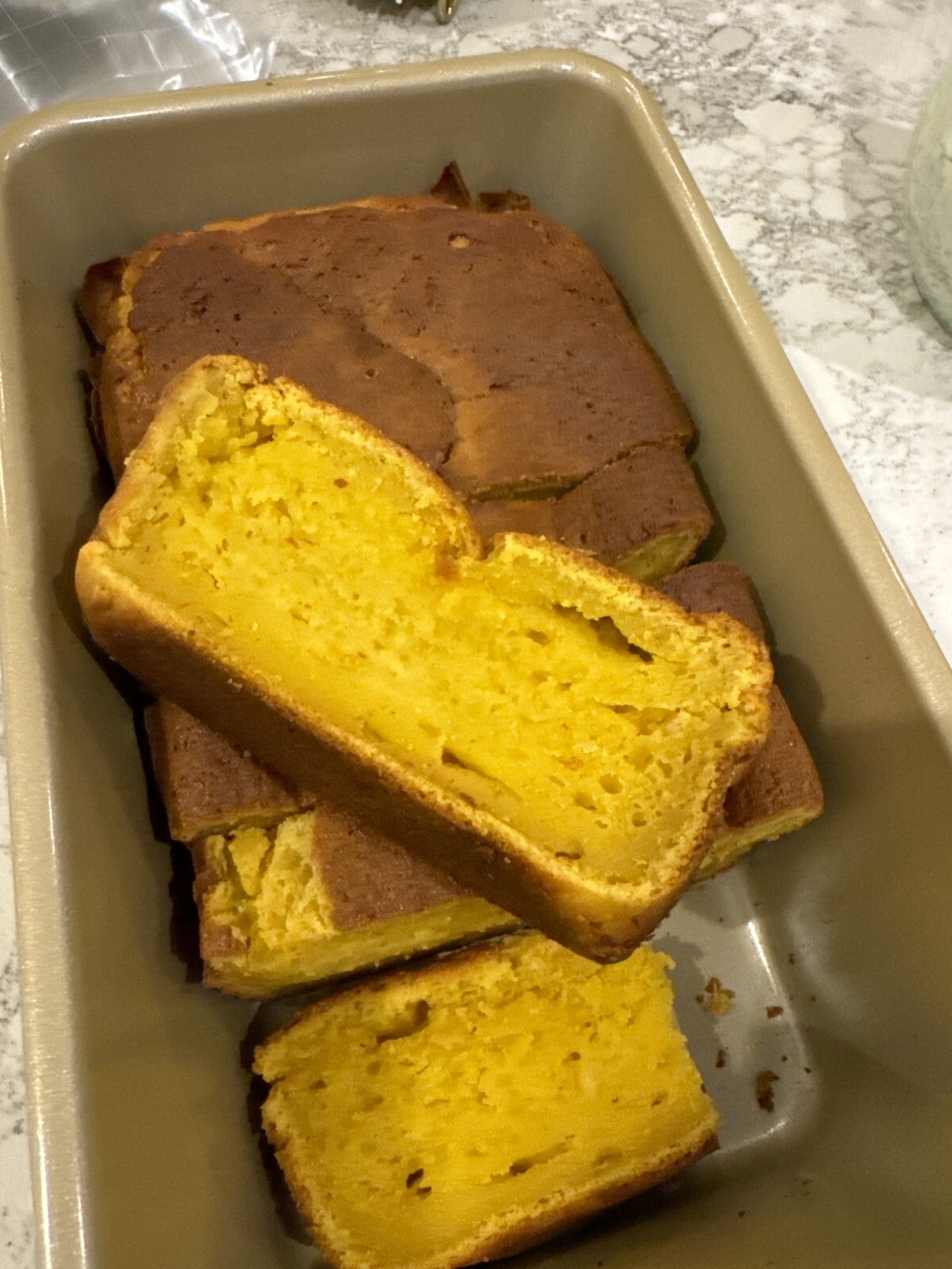 A loaf of golden pumpkin bread in a rectangular baking pan, with two thick slices cut and laid on top, showing a moist, dense texture reminiscent of Whole Orange Blender Cake. The slightly browned top sits on a marbled countertop.