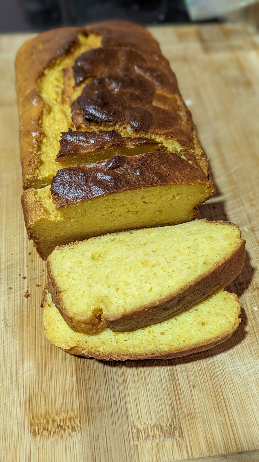A loaf of cornbread sits on a wooden cutting board, with two slices cut and laid in front of it. Its golden yellow hue and slightly browned top are reminiscent of the hearty color found in a Whole Orange Blender Cake.