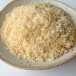 A close-up of a ceramic bowl filled with light-colored, flaky Italian breadcrumbs and panko on a white surface.