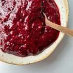 A close-up of a ceramic bowl filled with thick, chunky chia jam. A wooden spoon rests in the vibrant red spread, highlighting its glossy texture, visible seeds, and wholesome twist on classic raspberry jam.