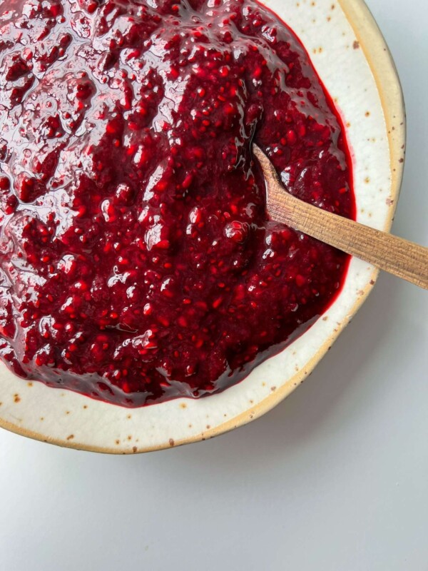 A close-up of a ceramic bowl filled with thick, chunky chia jam. A wooden spoon rests in the vibrant red spread, highlighting its glossy texture, visible seeds, and wholesome twist on classic raspberry jam.