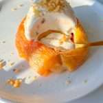 A classic dessert featuring baked apples topped with a scoop of vanilla ice cream and crushed nuts, elegantly served on a white plate with a spoon scooping into the warm, sweet treat.