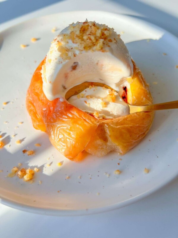 A classic dessert featuring baked apples topped with a scoop of vanilla ice cream and crushed nuts, elegantly served on a white plate with a spoon scooping into the warm, sweet treat.