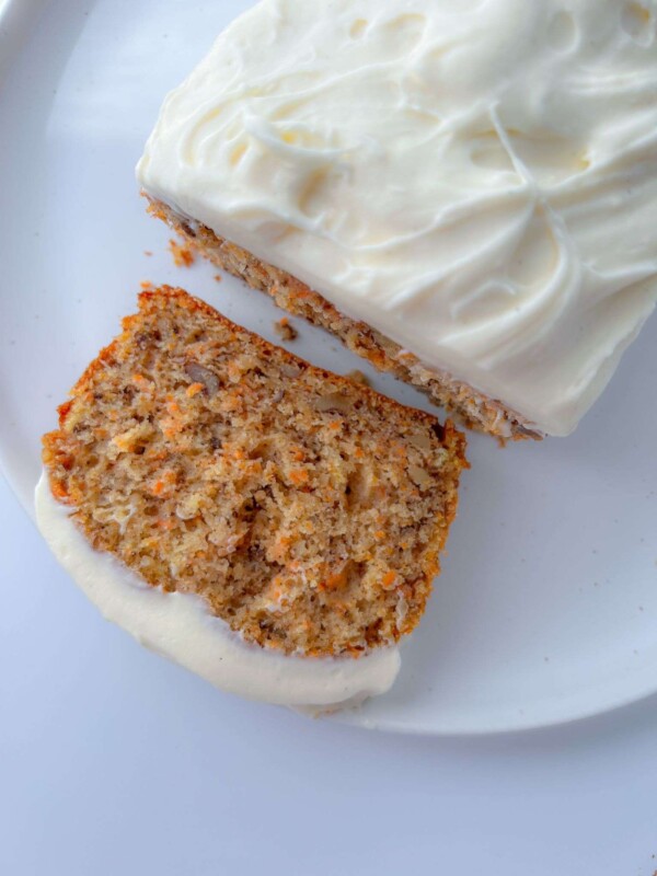 A close-up of an easy carrot cake with cream cheese frosting, with one slice cut and lying flat on a white plate. The moist carrot cake is speckled with orange carrot bits, making it look irresistible.