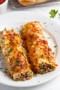 Two pieces of lasagna with crepes filled with minced meat and topped with tomato sauce and melted cheese, garnished with chopped parsley, served on a white plate.