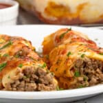 Two stuffed cannelloni filled with minced meat, topped with melted cheese and herbs, served on a white plate. A small ramekin of red sauce is in the background.