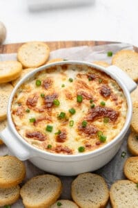 A white ceramic dish filled with baked dip topped with golden-brown cheese and chopped green onions. Slices of toasted baguette are arranged around the dish on a wooden board.