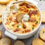 A white baking dish filled with creamy, bubbly artichoke and spinach dip topped with melted cheese and garnished with chopped green onions. A round, crispy bread slice is dipped into the dish, with more slices surrounding it.