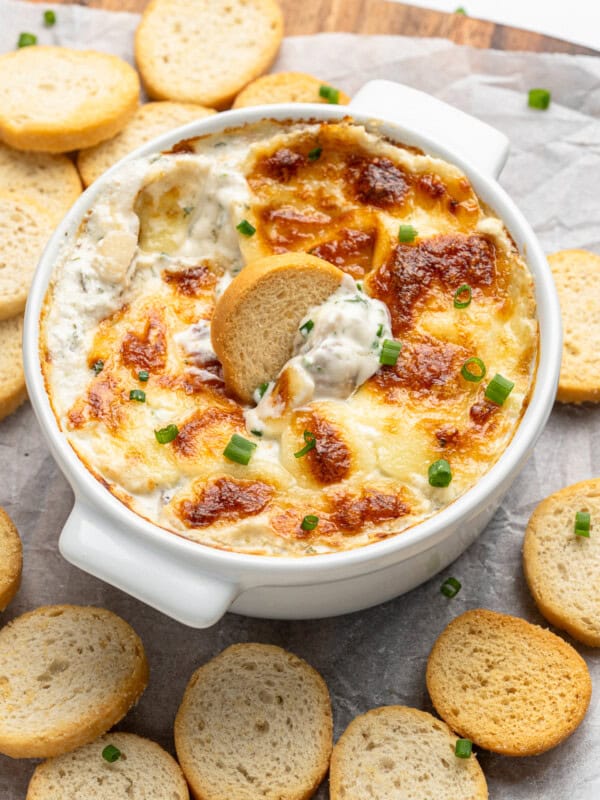 A white baking dish filled with creamy, bubbly artichoke and spinach dip topped with melted cheese and garnished with chopped green onions. A round, crispy bread slice is dipped into the dish, with more slices surrounding it.