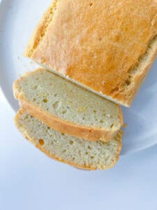 A loaf of golden-brown Healthy Sandwich Bread sits on a white plate, with two slices cut and placed in front, revealing a light and airy crumb texture.