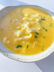 A close-up of a bowl of Hidden Veggie Soup featuring yellow chicken noodle soup with wide noodles, shredded chicken, and sprinkled fresh herbs, served in a light-colored bowl.