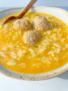 A bowl of Hidden Veggie Meatball Soup with yellow broth, rice, and three meatballs, served with a wooden spoon resting on the side.
