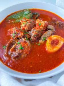 A white bowl filled with rich, red stew made from Authentic Italian Tomato Sauce, featuring chunks of beef, a bone with marrow, and garnished with chopped herbs and a bay leaf.
