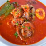 A bowl of rich, red stew with pieces of meat, a hard-boiled egg, a large bay leaf, and chopped herbs in an authentic Italian tomato sauce.