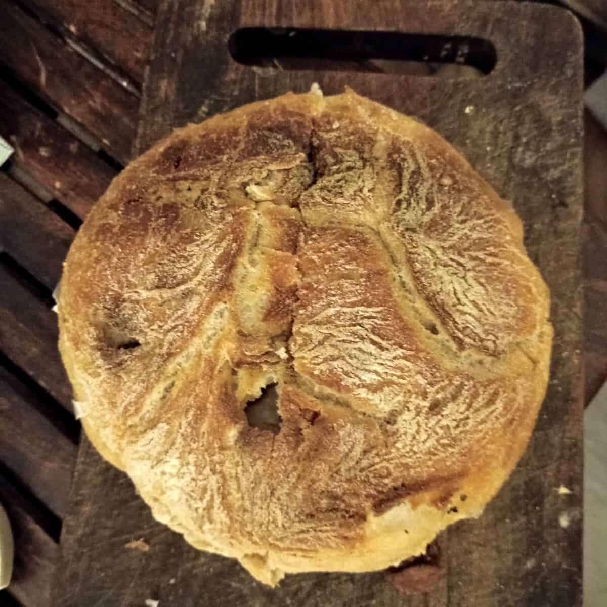 A round loaf of rustic bread with a golden-brown, textured crust sits on a dark wooden cutting board. The loaf has distinctive cracks and patterns on its top surface.