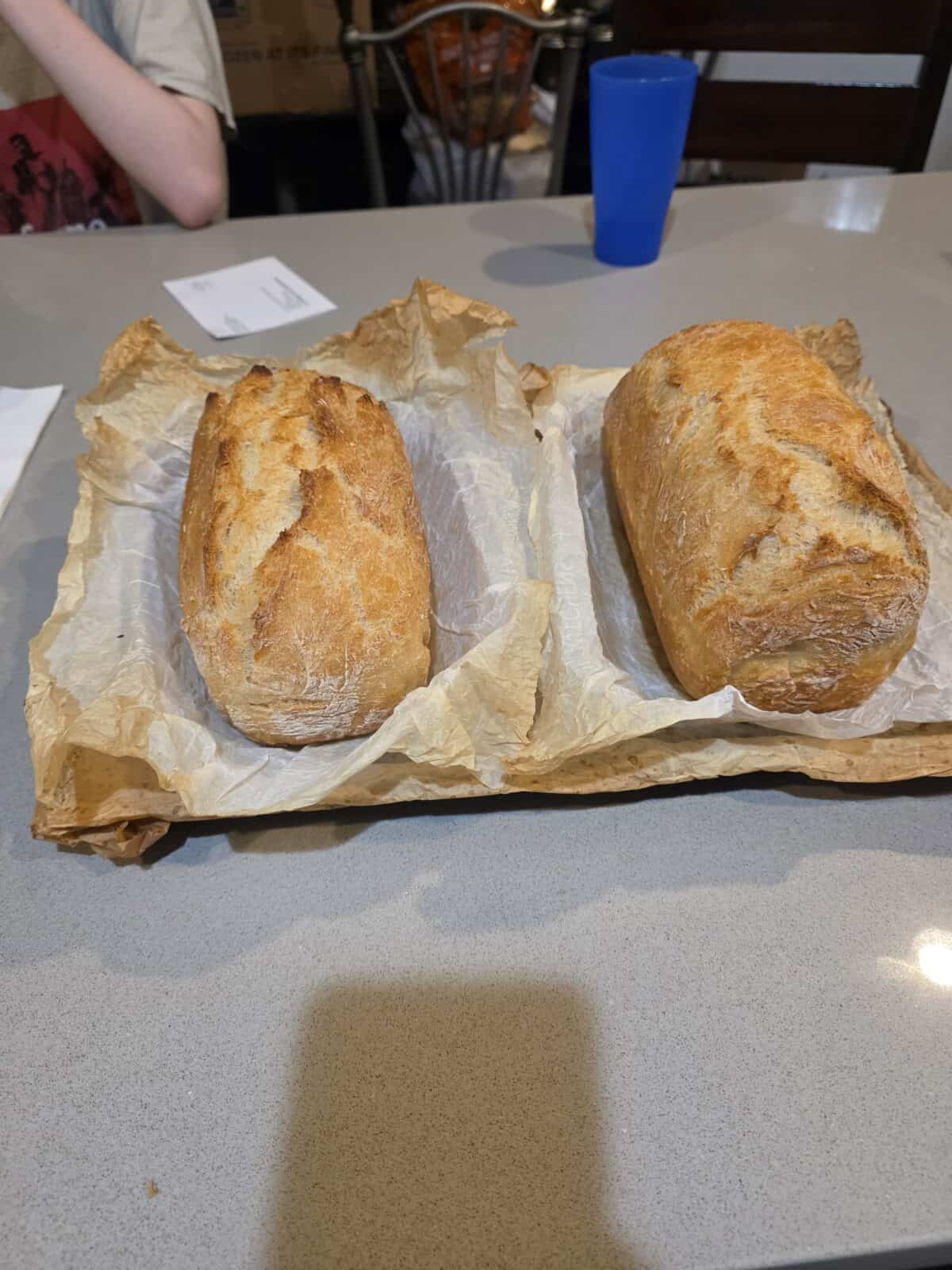 Two golden-brown loaves of No Knead Bread rest on parchment paper atop a baking sheet on a gray countertop. A child sits at the table in the background, along with a blue cup and papers.