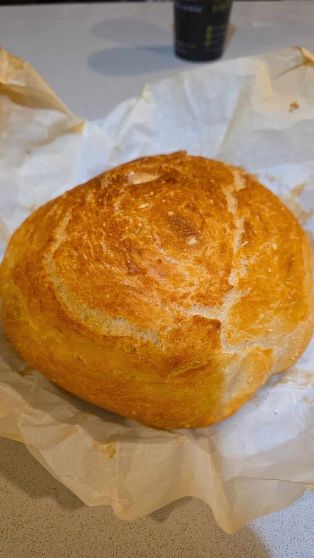 A round, golden-brown loaf of no knead bread sits on white parchment paper atop a light-colored countertop, with a blurred cup in the background.