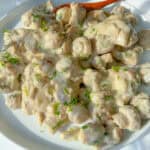 A plate of sliced mushrooms in a creamy white sauce, reminiscent of a Mushroom Chicken Skillet, is garnished with chopped fresh herbs and sits on a white surface with a wooden spoon beside the dish.
