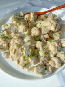 A plate of sliced mushrooms in a creamy white sauce, reminiscent of a Mushroom Chicken Skillet, is garnished with chopped fresh herbs and sits on a white surface with a wooden spoon beside the dish.