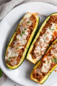 Three Stuffed Zucchini Boats filled with a hearty meat sauce, topped with melted cheese and chopped herbs, and elegantly served on a white plate.