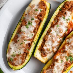 Three Stuffed Zucchini Boats filled with a hearty meat sauce, topped with melted cheese and chopped herbs, and elegantly served on a white plate.