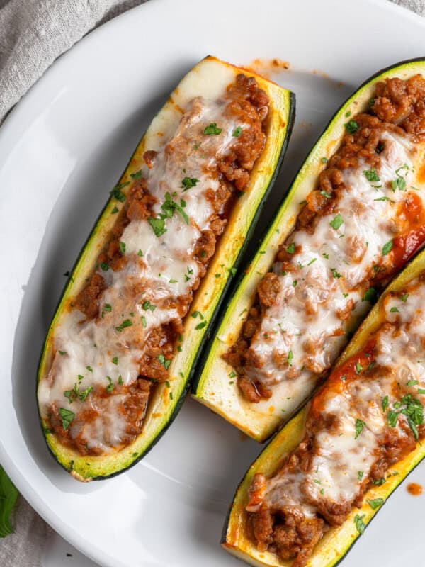 Three Stuffed Zucchini Boats filled with a hearty meat sauce, topped with melted cheese and chopped herbs, and elegantly served on a white plate.