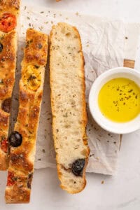 Slices of focaccia bread topped with olives and herbs on parchment paper, accompanied by a small white bowl of olive oil with herbs, set on a light marble surface.