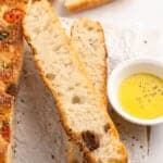 Slices of freshly baked focaccia bread with herbs and tomatoes are laid on parchment paper. A small bowl of olive oil is on the side for dipping.