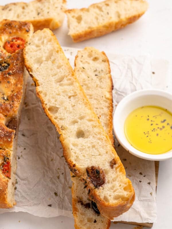 Slices of freshly baked focaccia bread with herbs and tomatoes are laid on parchment paper. A small bowl of olive oil is on the side for dipping.