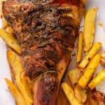 A roasted leg of lamb garnished with herbs is served on a wooden board alongside golden potato wedges and roasted garlic cloves.