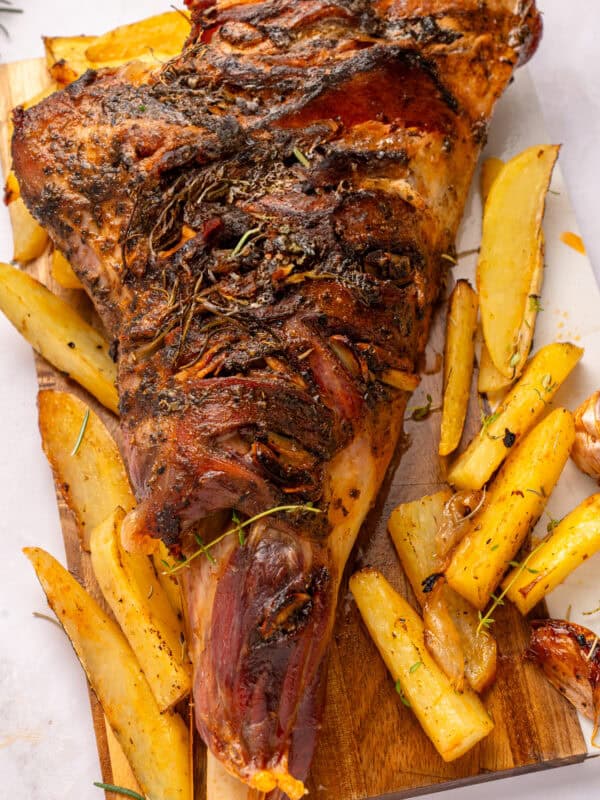 A roasted leg of lamb garnished with herbs is served on a wooden board alongside golden potato wedges and roasted garlic cloves.