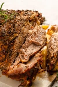 Close-up of a juicy, roasted lamb shoulder garnished with herbs. The meat is tender and shredded, with crispy, seasoned edges and visible sprigs of rosemary.