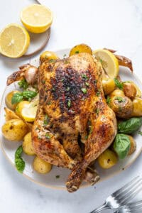A whole roasted chicken garnished with herbs sits alongside roasted baby potatoes, garlic bulbs, lemon halves, and fresh basil leaves on a white plate. No Knead Focaccia completes the meal, with two forks resting nearby on a marble surface.