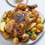 A roasted whole chicken garnished with herbs, surrounded by golden potatoes, garlic, and lemon wedges on a plate—perfect alongside slices of No Knead Focaccia. A halved lemon sits on a small plate in the background, with two forks in the foreground.