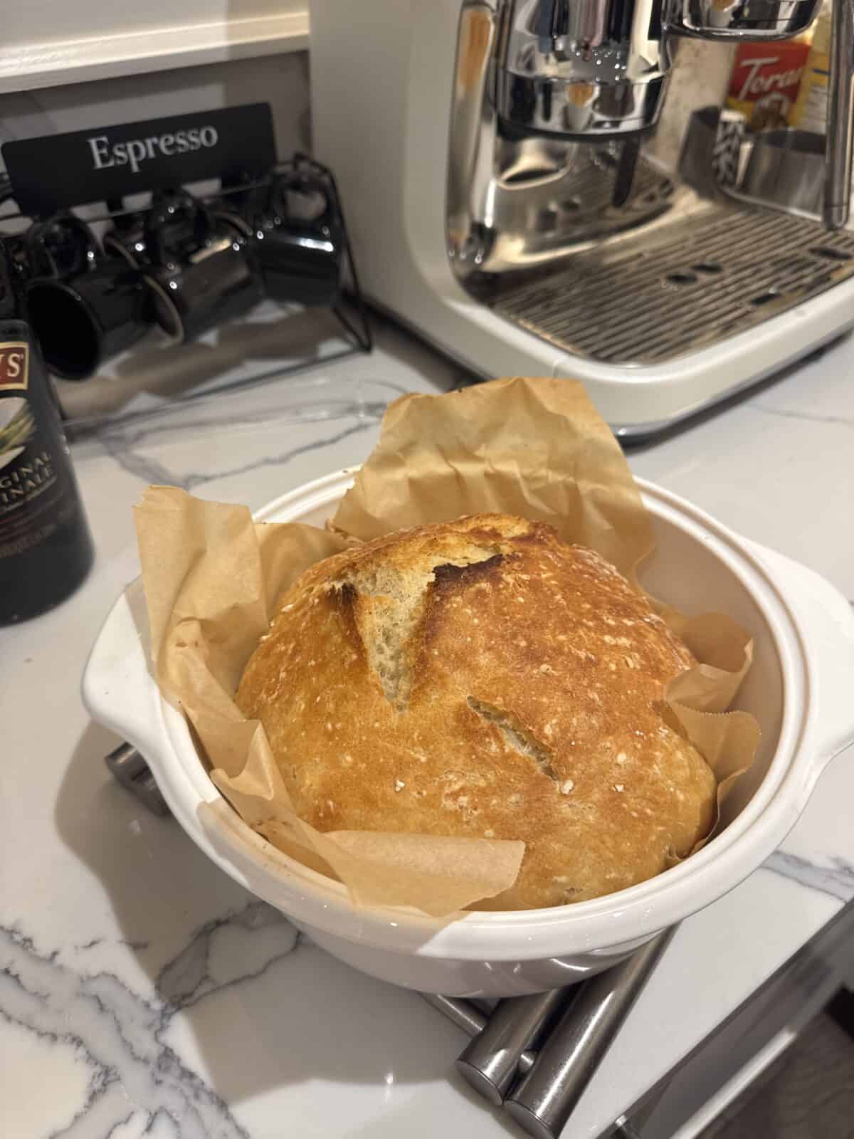 A round loaf of No Knead Bread with a golden-brown crust sits on parchment paper in a white ceramic dish on a marble countertop, next to an espresso machine and coffee mugs.