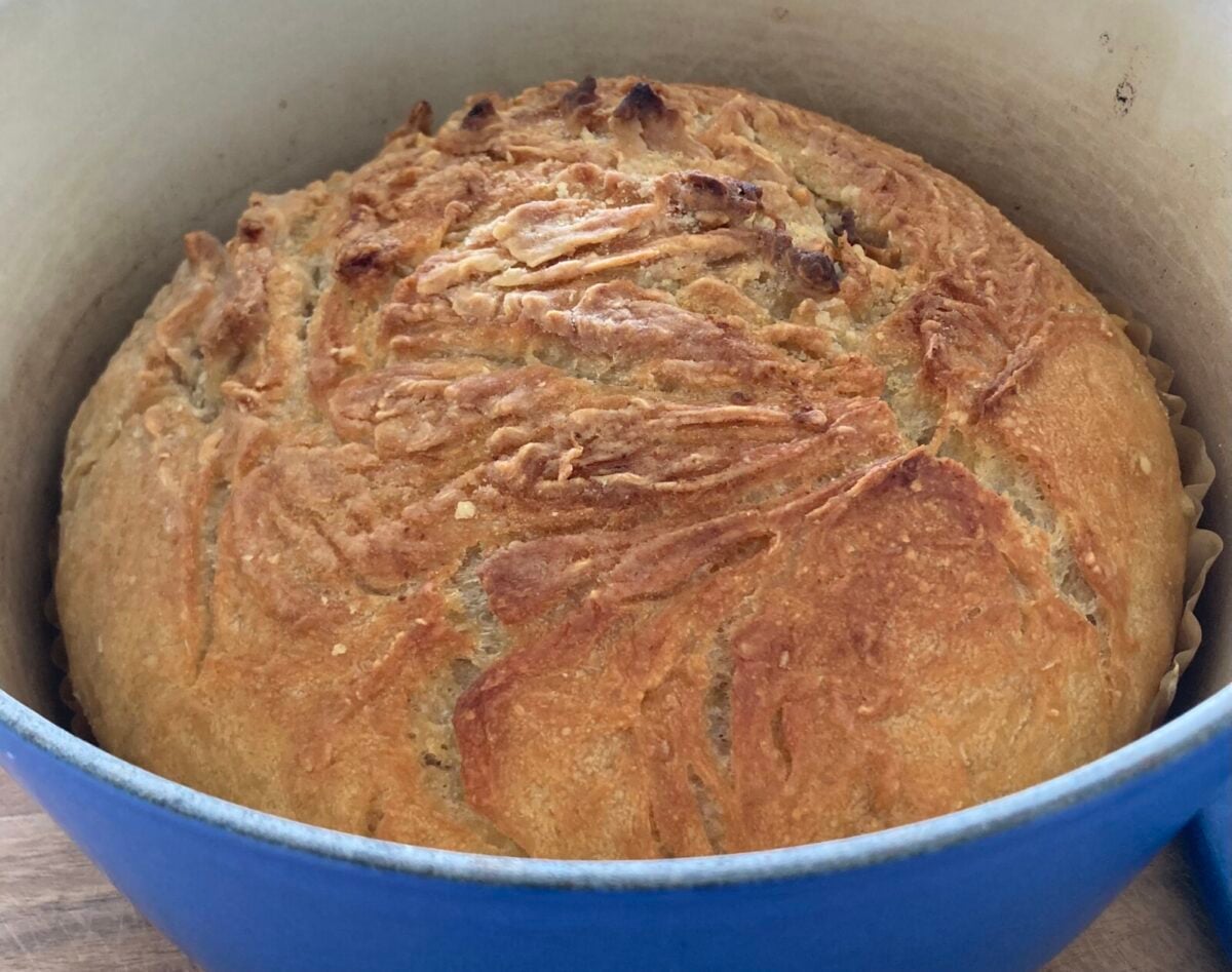 A round loaf of No Knead Bread sits in a blue Dutch oven, its crusty, golden-brown surface adorned with rustic swirls and cracks.