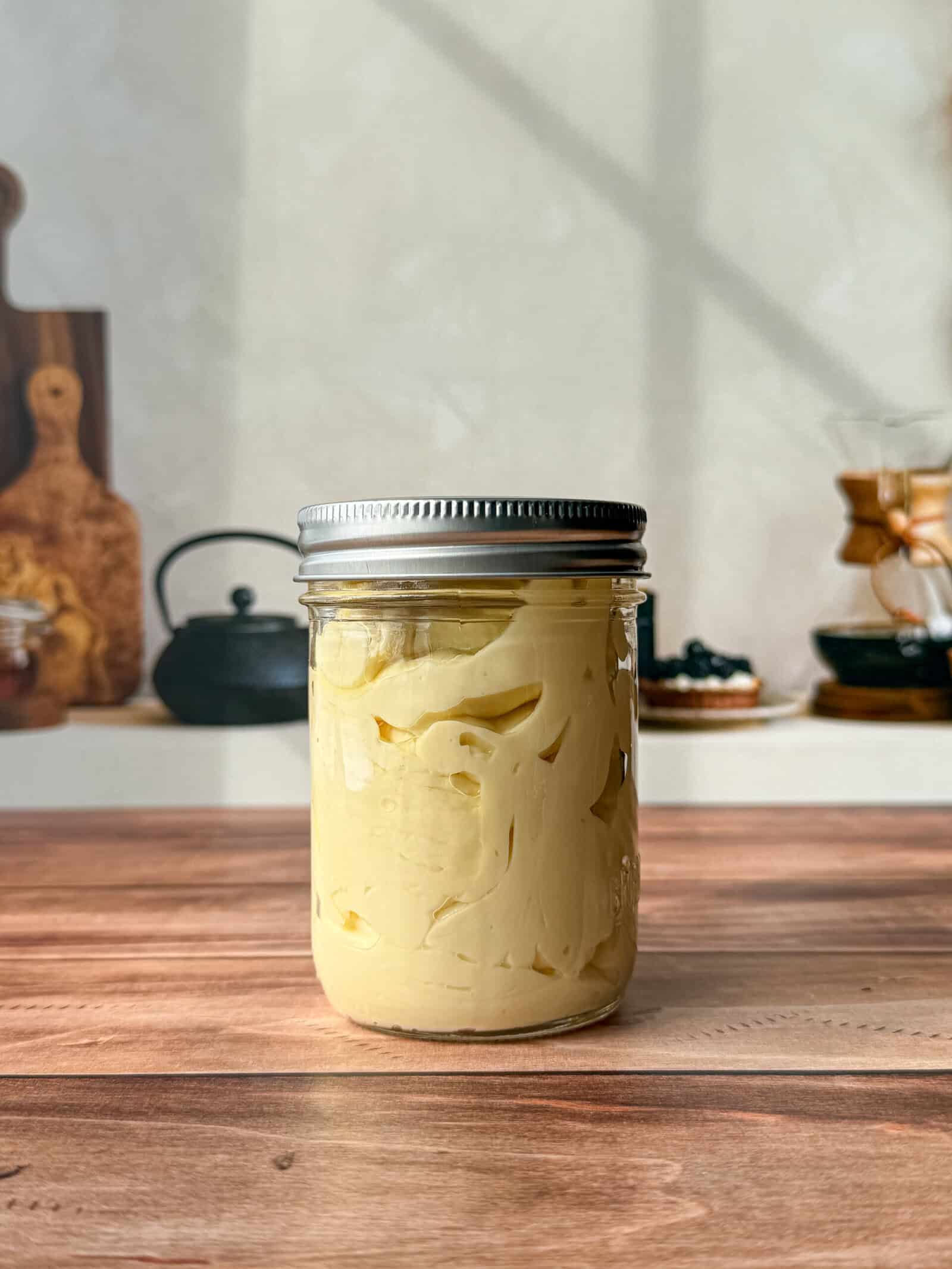 A glass jar with a metal lid filled with creamy yellow homemade mayo sits on a wooden surface in a kitchen, surrounded by blurred utensils and cookware in the background. - 6