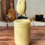 A spoon covered in creamy yellow homemade mayo is held above an open glass jar filled with mayonnaise, on a wooden table with kitchen items blurred in the background.