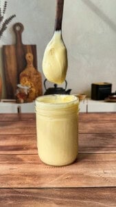 A spoon covered in creamy yellow homemade mayo is held above an open glass jar filled with mayonnaise, on a wooden table with kitchen items blurred in the background.