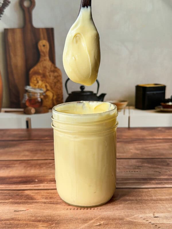 A spoon covered in creamy yellow homemade mayo is held above an open glass jar filled with mayonnaise, on a wooden table with kitchen items blurred in the background.