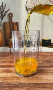 Olive oil is being poured from a measuring cup into a tall clear container with eggs, ready to be blended into homemade mayo, on a wooden table with kitchen utensils and decor in the background.