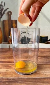 A hand cracks an egg into a clear measuring cup on a wooden counter, ready to make homemade mayo, with kitchen utensils and decor visible in the background.