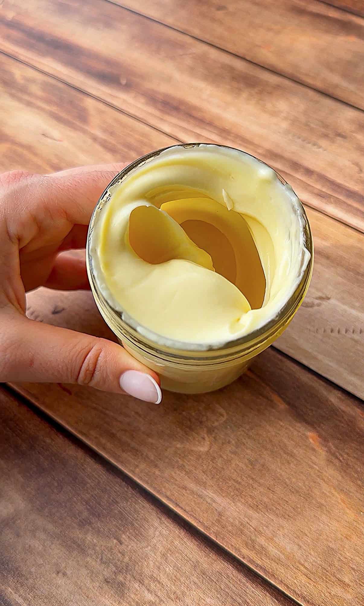 A hand holding a small glass jar filled with creamy, pale yellow homemade mayo on a wooden surface. The mayo inside has a smooth, swirled texture. - 5