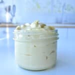 A glass jar filled with creamy, thick white homemade mayo sits on a glossy white surface in a bright kitchen. The background is softly blurred, showing kitchen utensils and a sink.